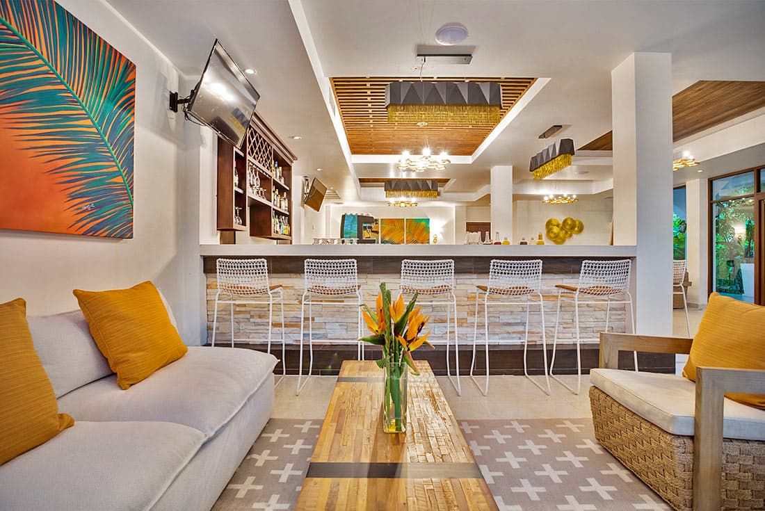 A modern hotel lobby with a high ceiling and wooden accents. Comfortable seating arrangements, including sofas and armchairs with orange cushions, are arranged around a central coffee table. A bar area with a counter and stools is visible in the background. Large windows offer natural light and views of the surrounding area. A colorful painting hangs on the wall, and a vase of flowers sits on the coffee table.