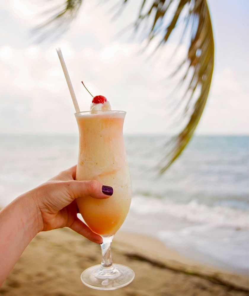 A hand holding a tropical drink with a cherry topping and a straw, set against a beach background.