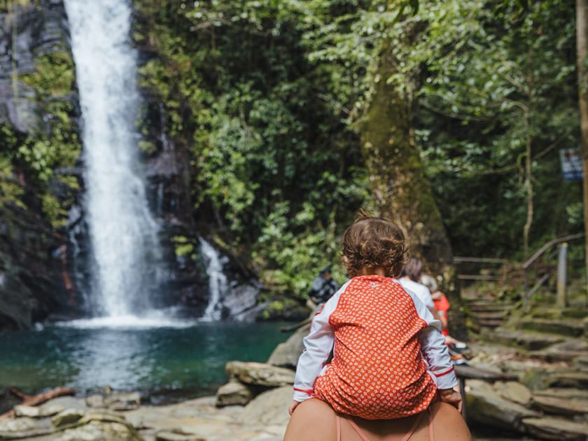 A child in a red polka dot outfit sits atop an adult's shoulders, admiring a scenic waterfall surrounded by lush greenery. A child in a red polka dot outfit sits atop an adult's shoulders, admiring a scenic waterfall surrounded by lush greenery.
