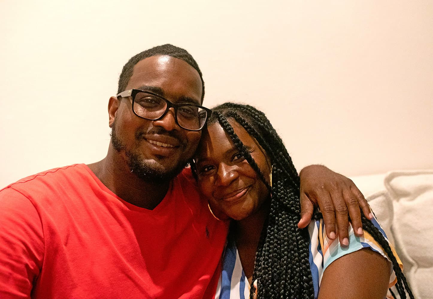 A man in an orange shirt and black glasses and a woman in a striped top embrace with smiles.