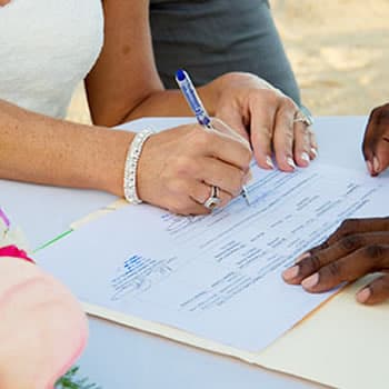 A bride in a white dress is signing a wedding document. The groom's hand is resting on the paper beside hers.