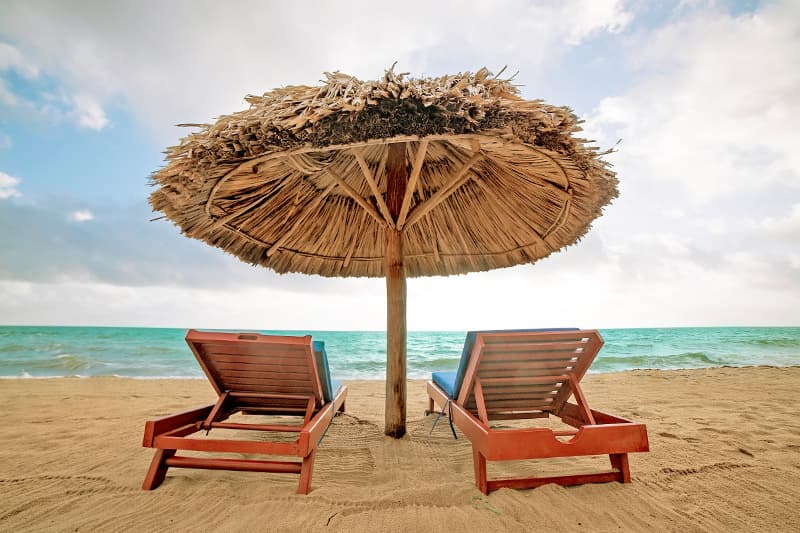 Two beach loungers under a thatched umbrella overlooking the ocean.