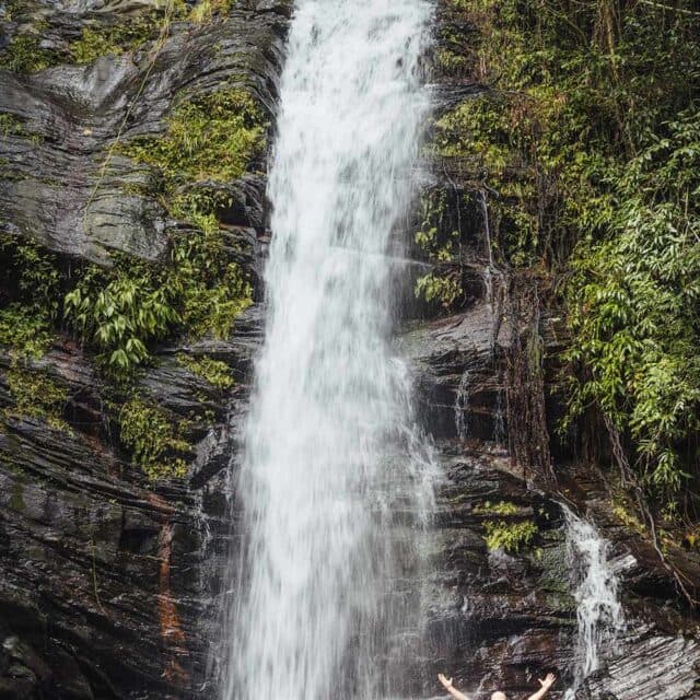 A man stands in a natural pool at the base of a tall, cascading waterfall. The surrounding area is lush with green vegetation.