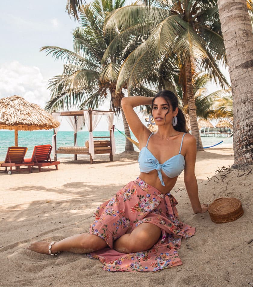 A woman in a blue top and floral pink skirt sits on the beach surrounded by palm trees and loungers.