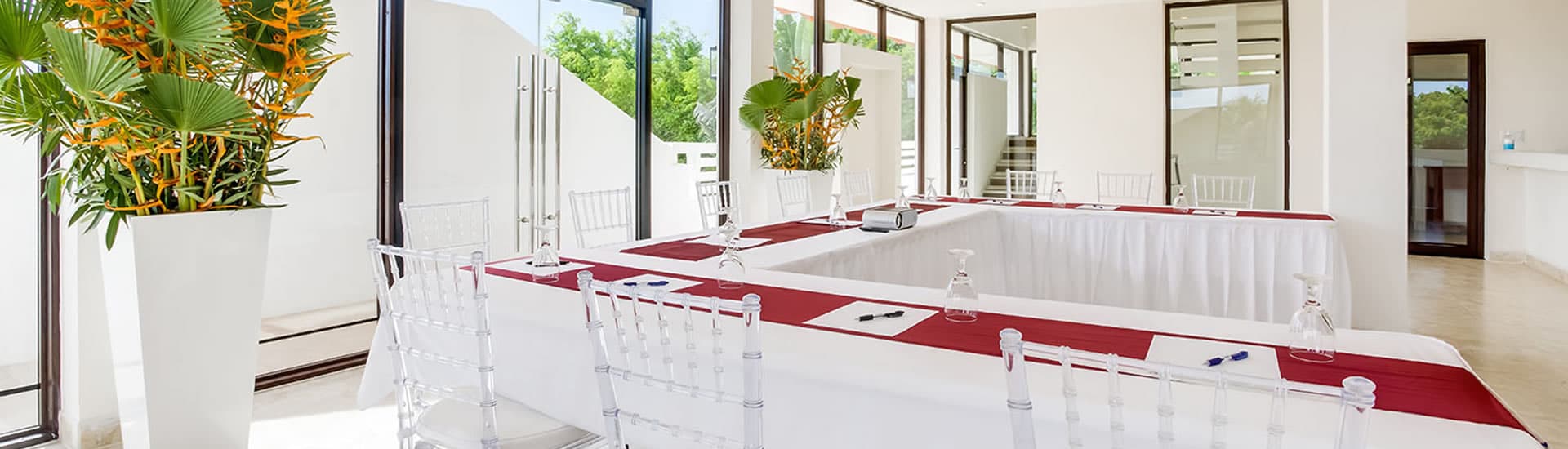 A modern meeting room with large windows overlooking a tropical garden. A long, white table with red runners is arranged in a U-shape, surrounded by clear ghost chairs. There are potted plants in the corners of the room.