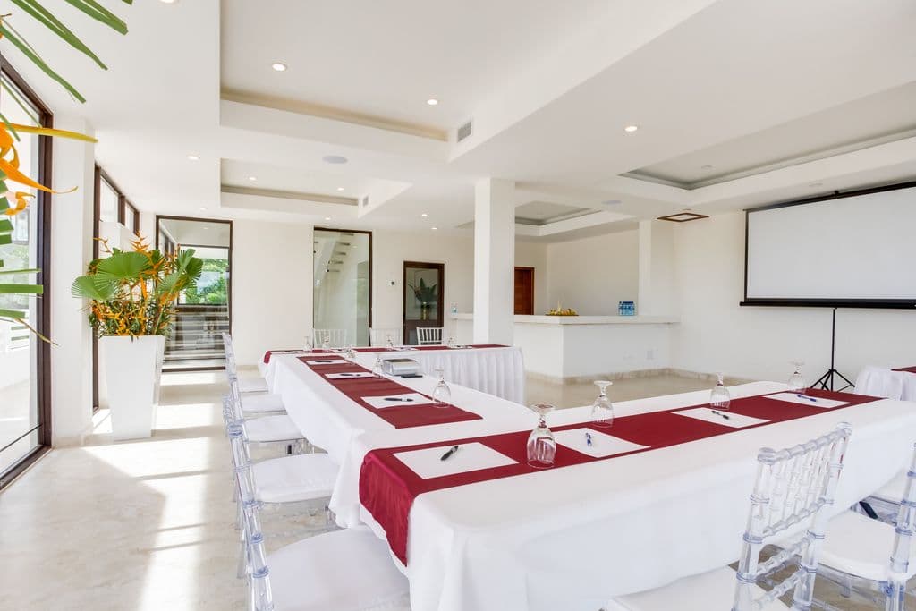 A spacious and modern meeting room with a U-shaped table arrangement. The room has a white tiled floor, white walls, and a high ceiling with exposed beams. There is a large palm tree in the corner, and a projection screen and projector at the front of the room. The table is covered in white tablecloths and has red runners. Clear plastic chairs are arranged around the table. There is a bar area with counter stools in the background.