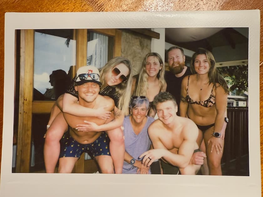 A group of six people poses together in swimwear, smiling and enjoying a sunny day by a window.