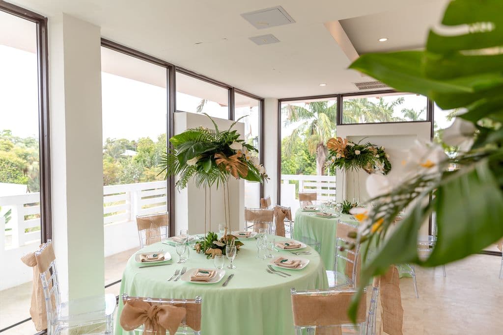 A beautifully decorated tropical wedding reception venue. Round tables are covered in light green tablecloths and adorned with floral centerpieces featuring orchids, palm leaves, and gold accents. Large windows offer views of lush greenery.