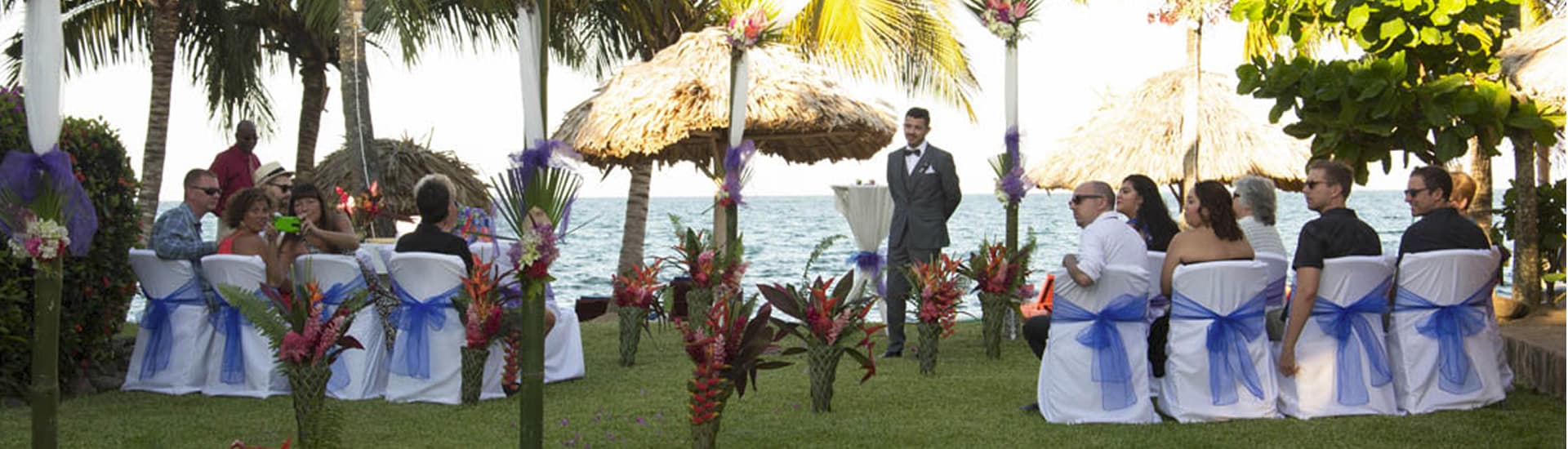 A groom waits for his bride at a wedding arch while the guests turn to look.