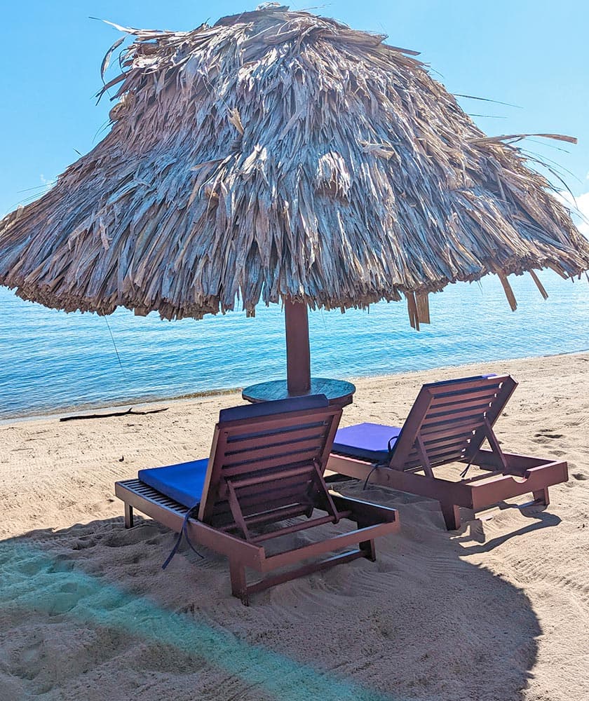 Two lounge chairs sit beneath a thatched umbrella on a sandy beach by the water.
