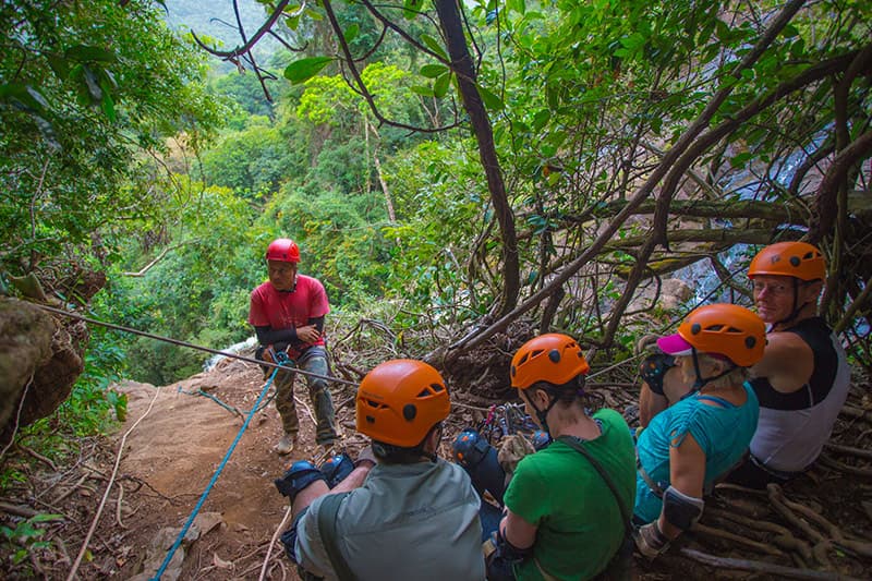 A group of climbers in helmets listens to an instructor on a steep, wooded slope.