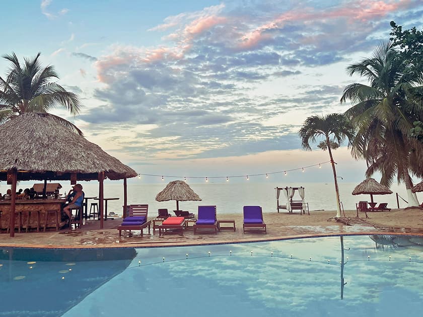 A tranquil beachfront resort scene with lounge chairs, a pool, and palm trees under a colorful sky.
