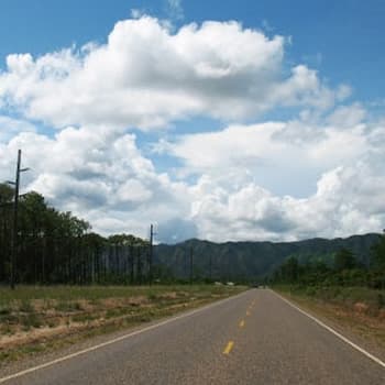 A long, straight road stretches into the distance under a blue sky with fluffy white clouds. Trees line the sides of the road, and mountains can be seen in the background.