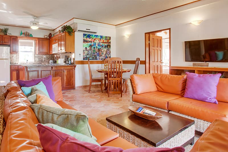 A bright living area featuring orange couches with colorful cushions, a wooden dining table, and a kitchen in the background.