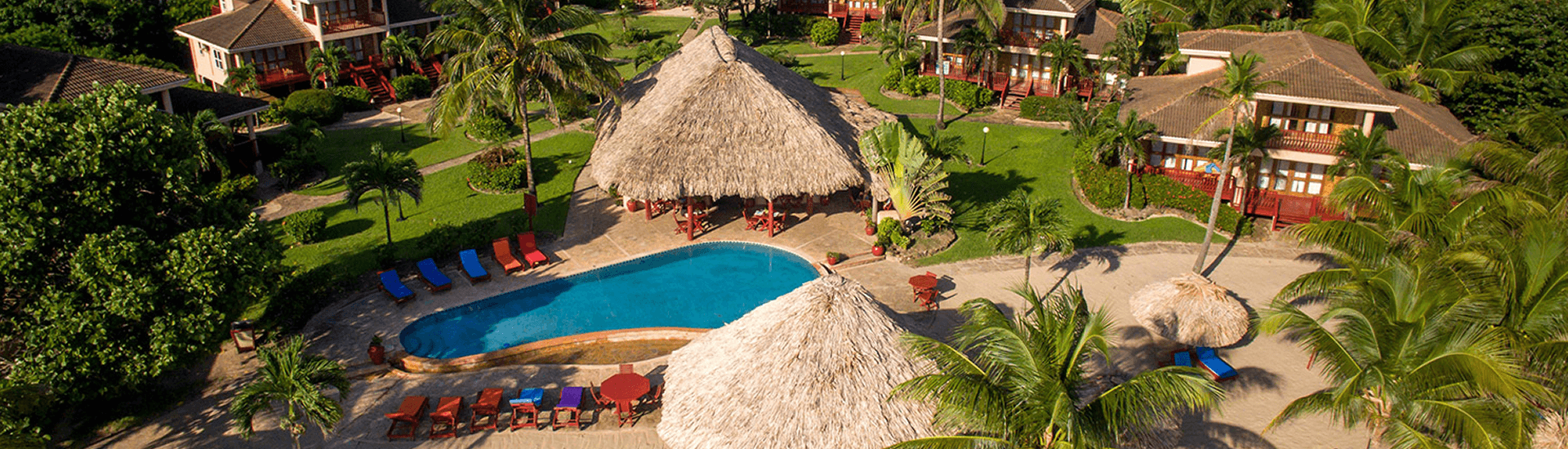 An aerial view of a tropical resort with a pool, thatched-roof structures, and colorful buildings.