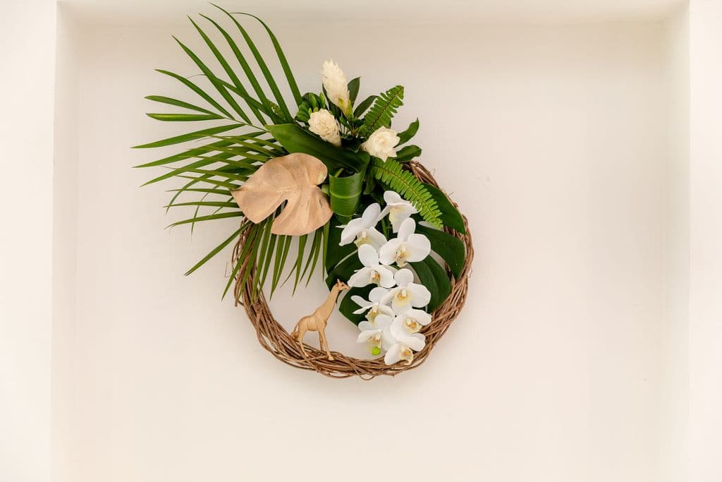 A circular wreath made of natural materials with a gold giraffe figurine hanging from it. The wreath is adorned with white orchids, palm leaves, and other tropical greenery.