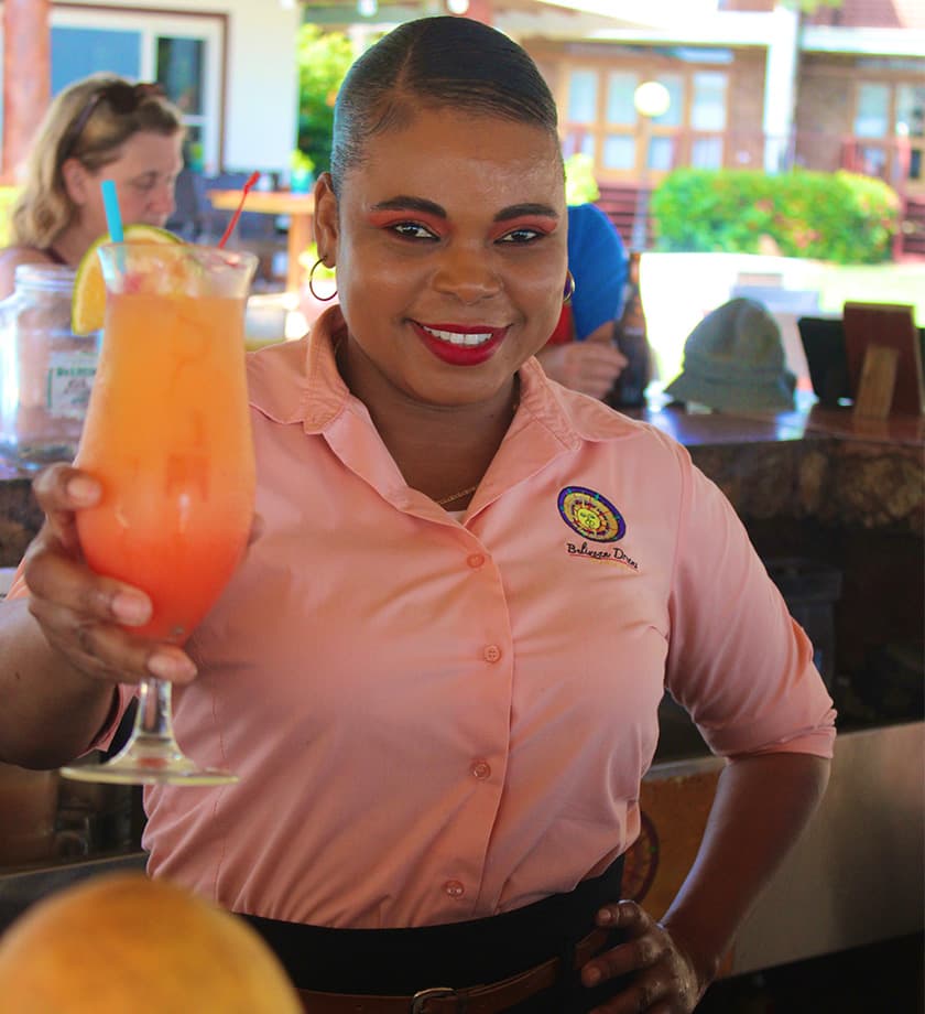A smiling woman in a pink shirt holds a colorful cocktail in a sunny bar setting.