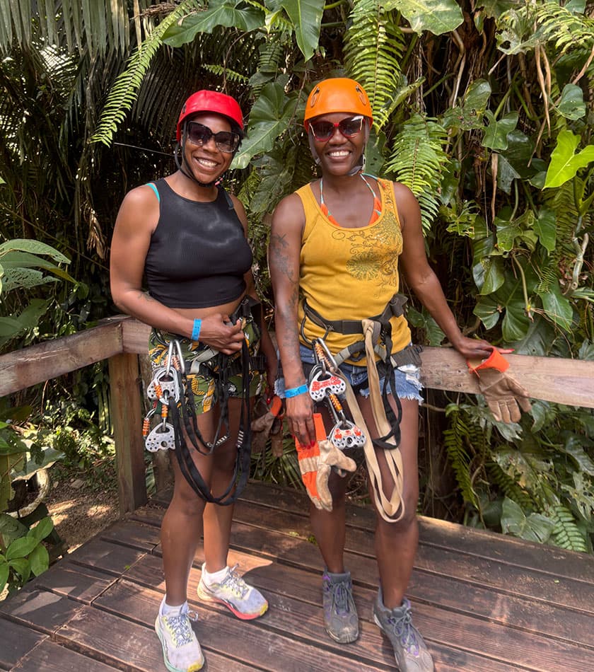 Two women wearing climbing gear and helmets smile together in a lush, green jungle setting.
