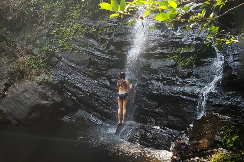 A person stands beneath a waterfall in a lush, rocky landscape.