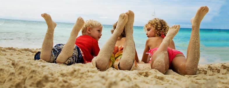 Three children sit on a sandy beach, facing the water with their sandy legs raised.