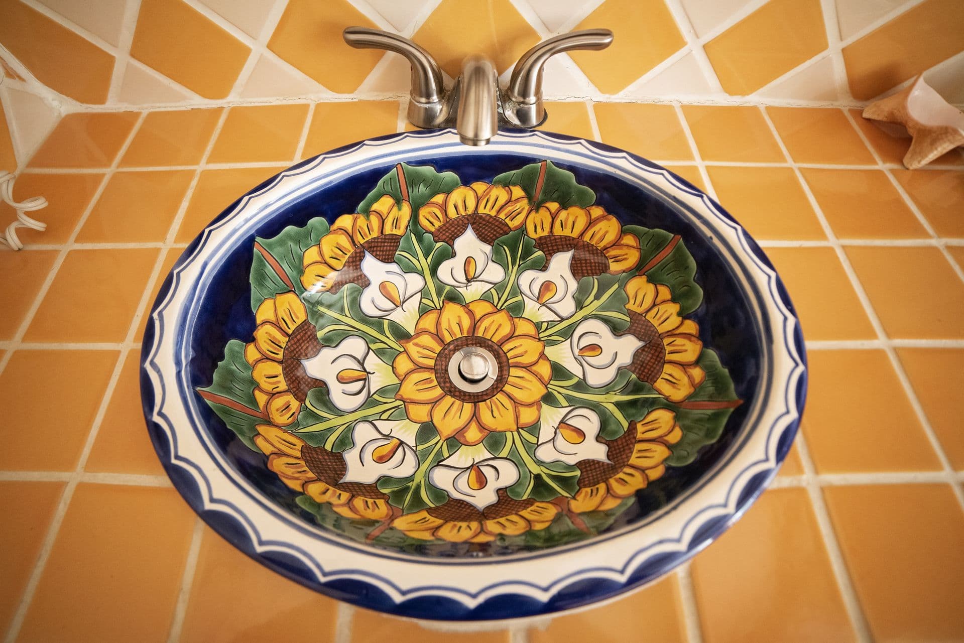 A circular ceramic sink with a colorful design featuring sunflowers, lilies, and green leaves. The sink is set against a backdrop of yellow and orange square tiles.
