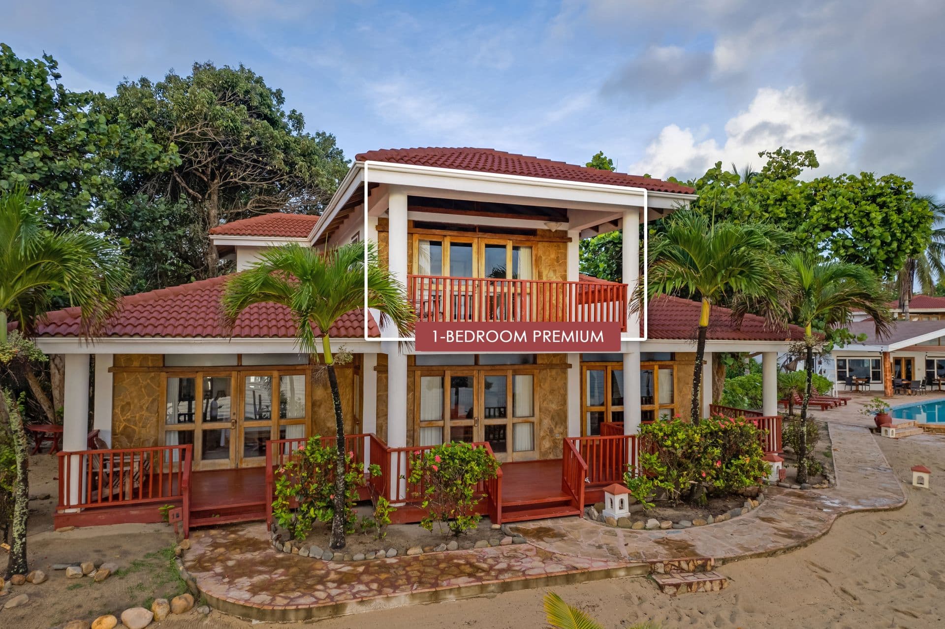 The image shows a 1-bedroom premium room at Belizean Dreams Resort in Hopkins Bay, Belize. The room is a two-story building with a red tile roof and a large balcony on the second floor. The room is surrounded by palm trees and lush greenery.