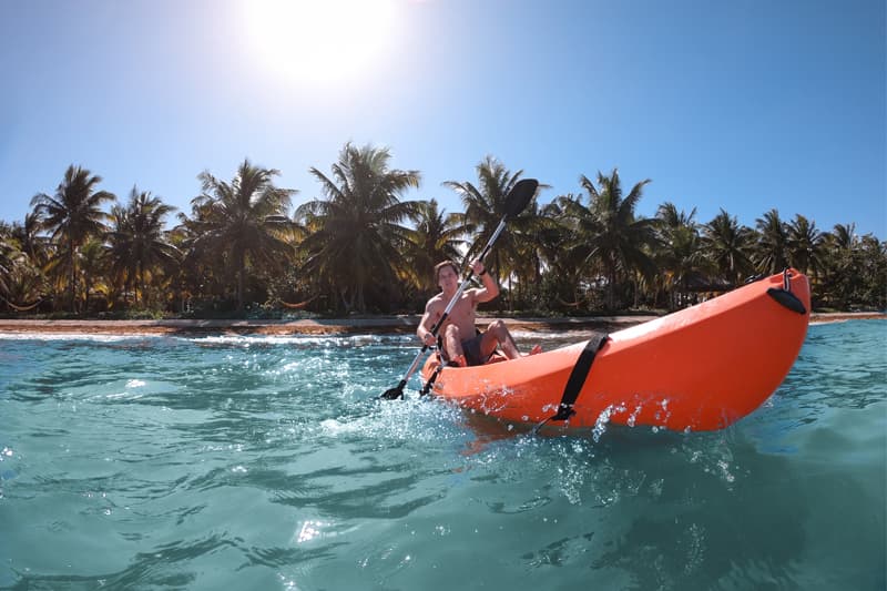 A person kayaking in bright orange attire on clear blue water with palm trees in the background.
