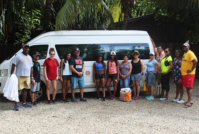 A diverse group of people poses together in front of a white van surrounded by lush greenery.