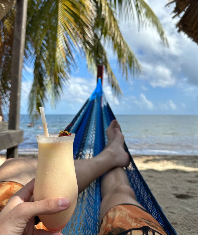 A person lounging in a hammock by the beach, holding a tropical drink with palm trees and water in the background.