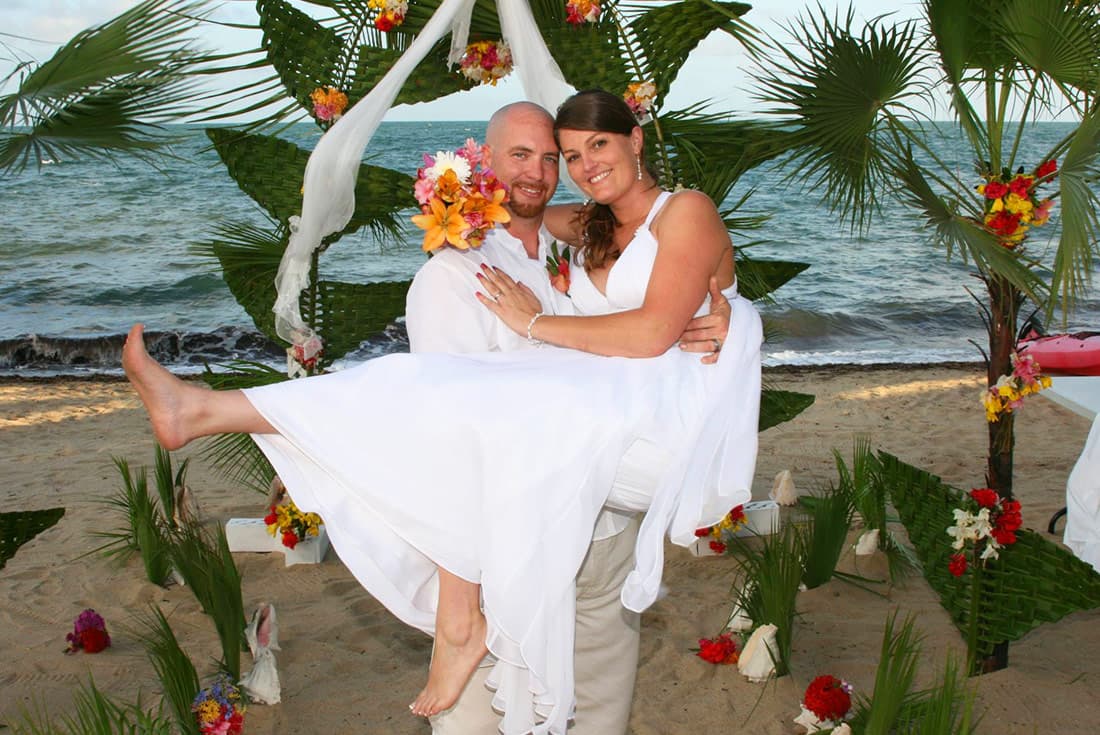 A happy groom carries his new bride on the beach in front of a lovely wedding arch made of tropical flowers in red and yellow.
