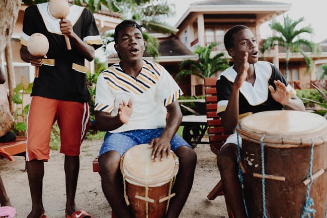 Three young men play traditional drums and instruments outdoors.