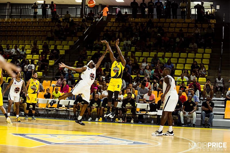 A basketball player in a yellow uniform shoots for the basket while a defender in white attempts to block the shot, with a crowd watching in the background. A basketball player in a yellow uniform shoots for the basket while a defender in white attempts to block the shot, with a crowd watching in the background.