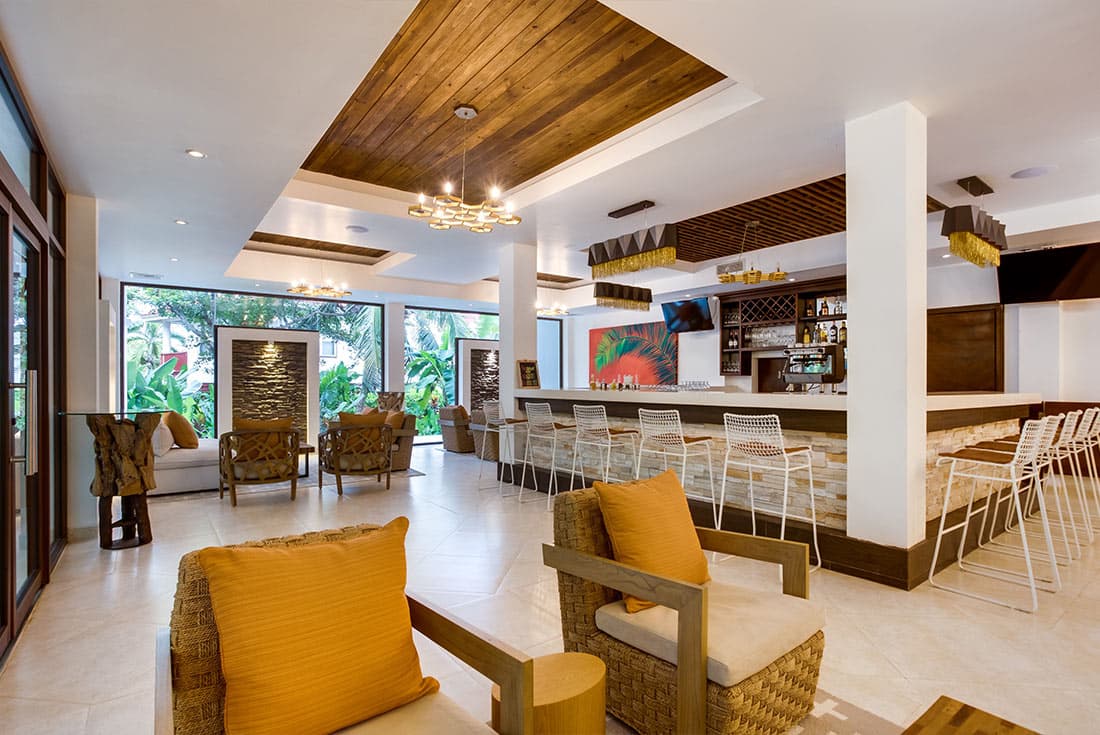 A spacious and modern hotel lobby with a high ceiling and wooden accents. Comfortable seating arrangements, including sofas and armchairs with orange cushions, are arranged around a central coffee table. A bar area with a counter and stools is visible in the background. Large windows offer natural light and views of the surrounding area.
