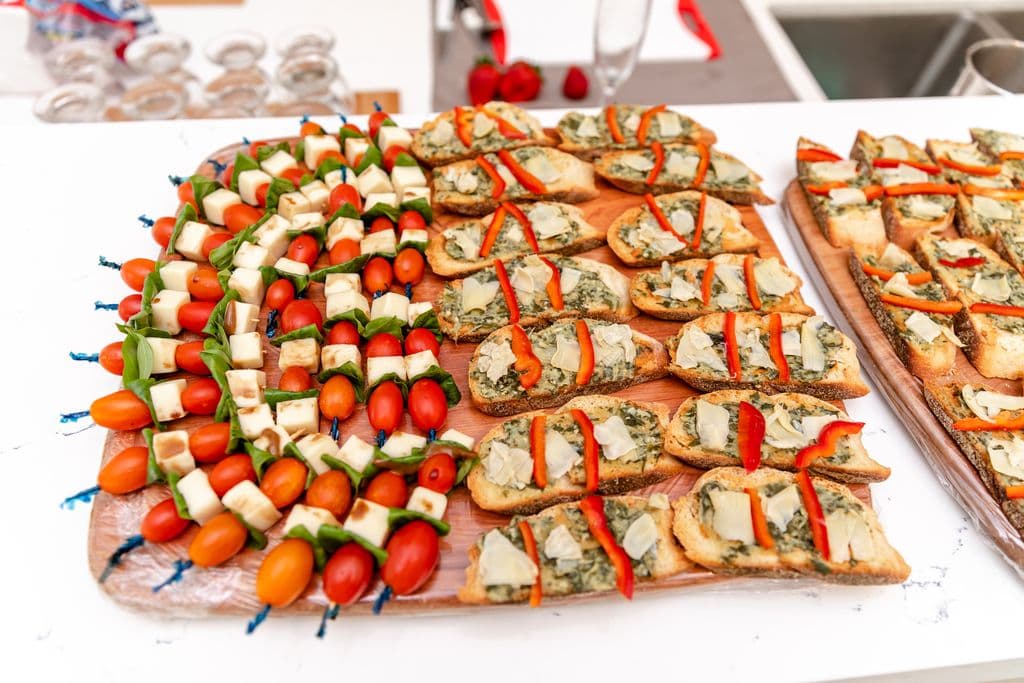 A platter of assorted appetizers, including skewers with grilled chicken, cheese, and cherry tomatoes, and open-faced sandwiches with pesto, red peppers, and cheese.