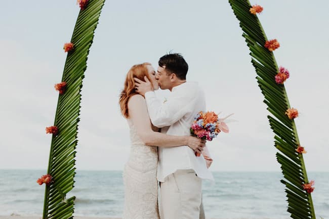 A couple kisses under a floral arch by the beach.