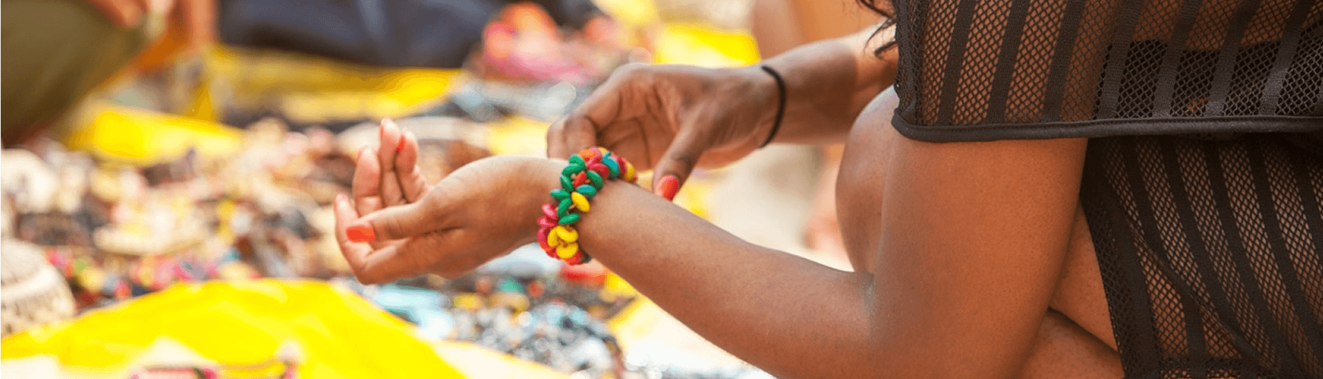 A woman is sitting at a market stall and looking at a colorful beaded bracelet on her wrist.