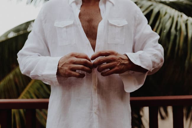 A person buttoning a white linen shirt stands in front of palm leaves.