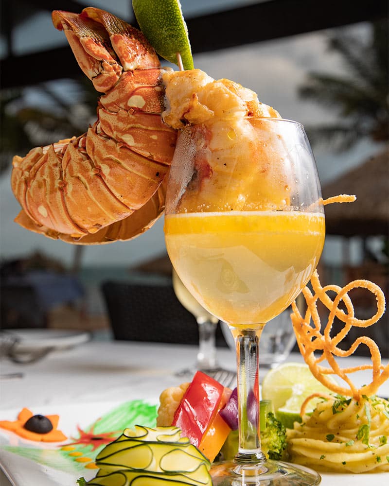A delicious-looking lobster dish served on a white plate. The lobster is cooked and seasoned, with a lime wedge perched on top. A glass of a golden-colored cocktail sits next to the lobster, and a colorful assortment of vegetables and pasta accompanies the dish.
