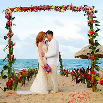 Man and woman dressed in wedding clothes, under flower arbor on beach, with ocean and blue sky in background.