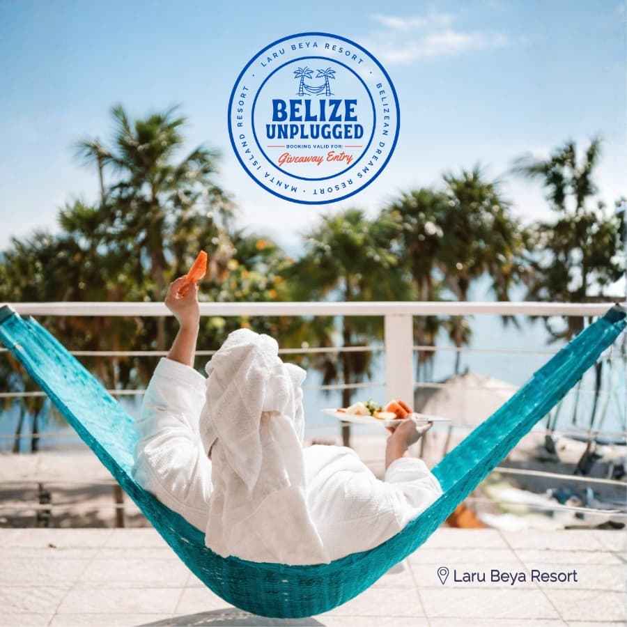 A person in a white robe relaxes in a hammock with a plate of food, overlooking a scenic ocean view at Laru Beya Resort.