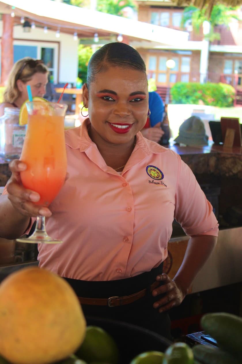 A smiling bartender in a pink shirt holds a colorful cocktail in front of a vibrant bar setting.