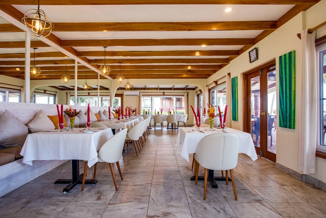 A spacious and modern restaurant with a high ceiling and exposed wooden beams. White tablecloths and chairs are arranged throughout the room, with a cushioned bench seating area along one side.