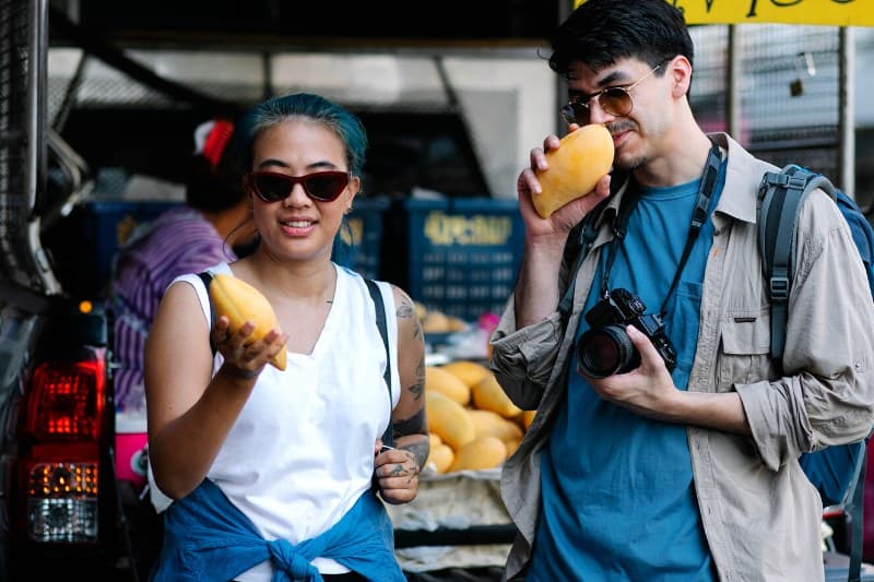 Two people hold mangoes while standing in a market, one wearing sunglasses and the other with a camera hanging from his neck.