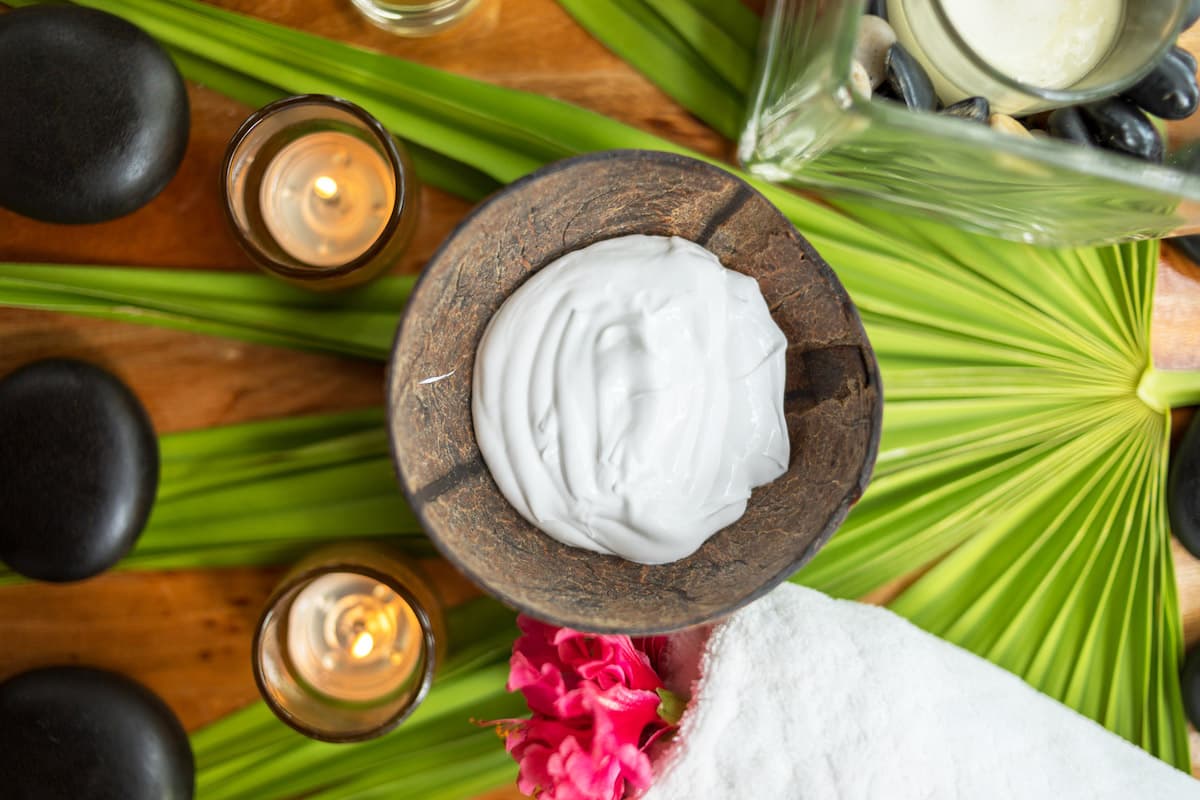 A relaxing spa scene with a coconut cream bowl, palm leaves, black stones, lit candles, and a white towel.