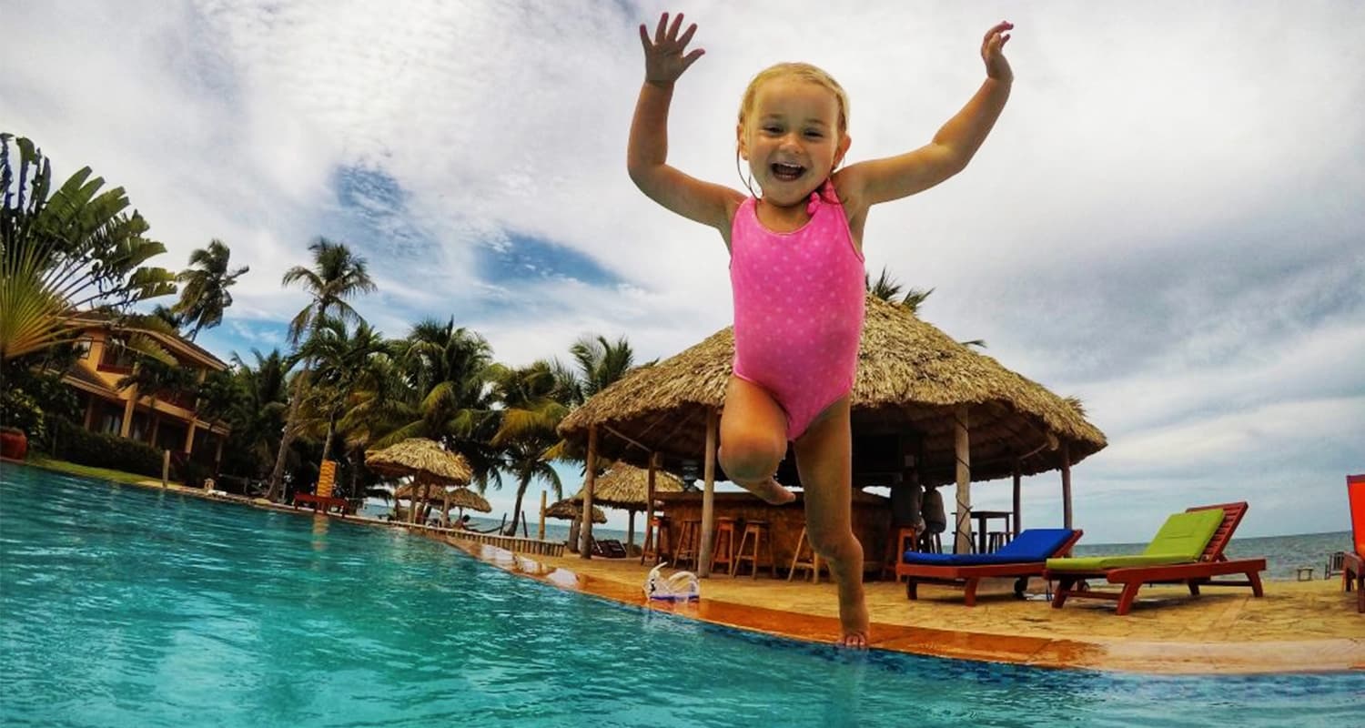 Toddler in pink bathing suit with arms in the air jumping into pool, near waters edge, with grass hut and lounge chairs in the background.