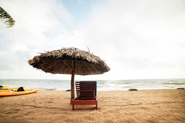 A beach scene featuring a wooden lounge chair under a thatched umbrella, with the ocean in the background.