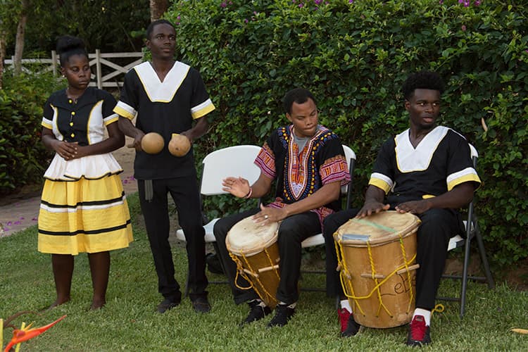 A group of four musicians, dressed in traditional attire, perform with drums and maracas in a garden setting.