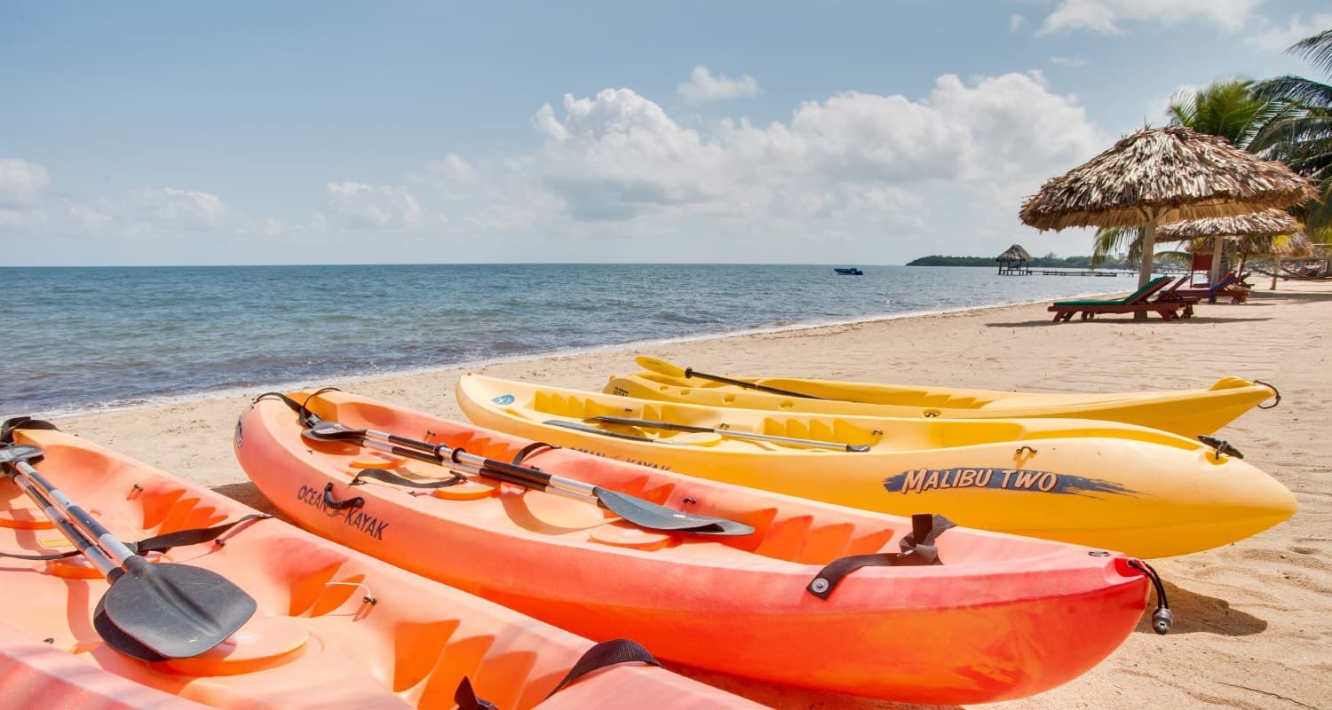 Several kayaks lined up on a sandy beach with a clear blue ocean in the background. The kayaks are bright orange and yellow, and there are palm trees and lounge chairs visible on the beach