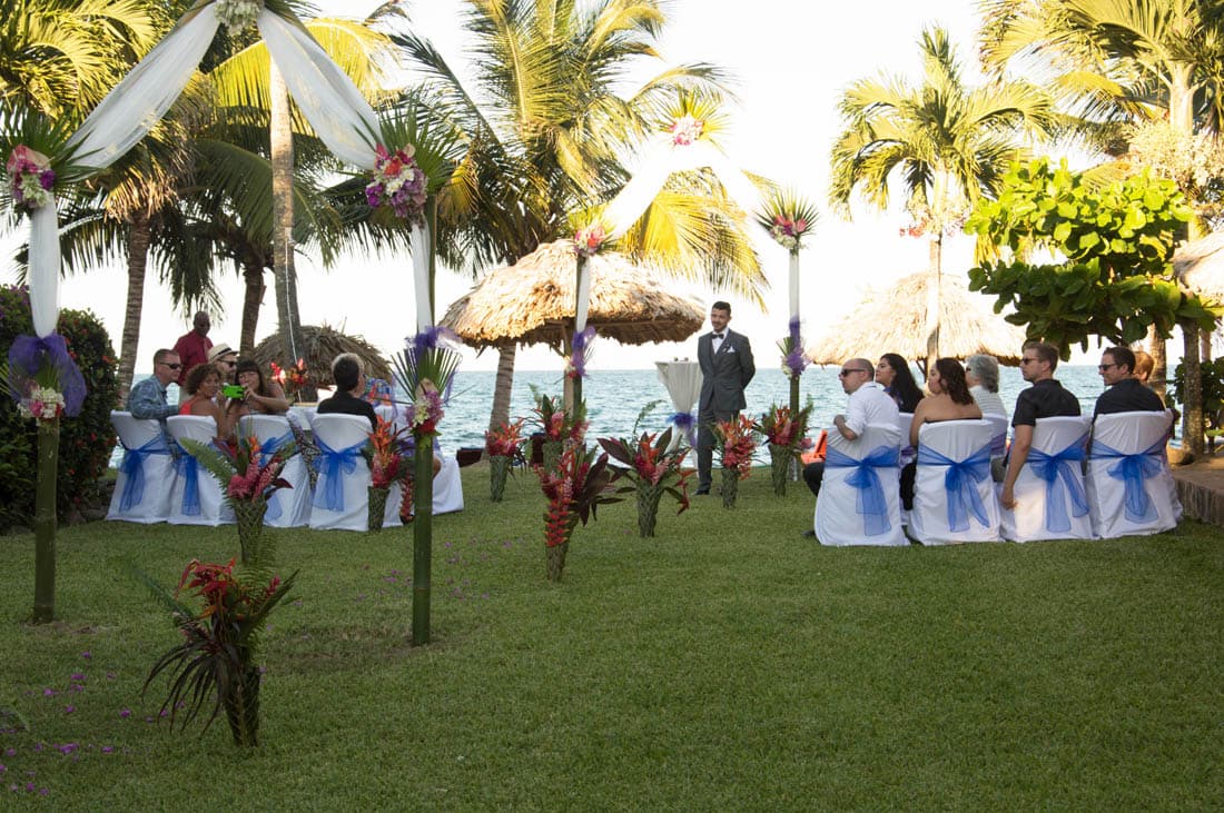 A groom stands waiting for his bride at an outdoor tropical beach wedding, surrounded by guests in chairs with white covers and blue bows.