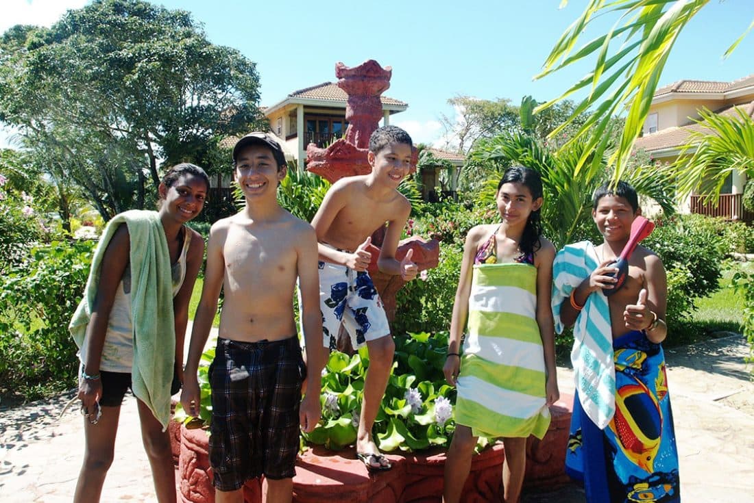 A group of teens in swimwear poses happily around a red stone fountain surrounded by jungle foliage.